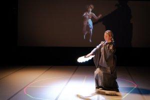 A photo of this event's speaker, Yasuko Yokoshi, performing a dance piece on a stage wearing a kimono and holding a fan.