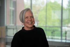 Professor Natalie Gummer smiles at the camera, wearing a black sweater and silver hoop earrings, with a softly blurred background of large windows and green trees.