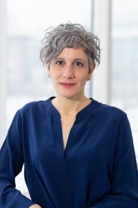 Lori Khatchadourian poses for a professional portrait wearing a navy blue blouse, with short gray hair and a softly blurred, bright background.