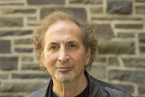 Peter Balakian poses for a portrait in front of a stone wall, wearing a black shirt and black leather jacket.