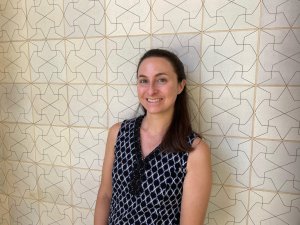 Jennifer Manoukian smiles for a portrait in front of a cream-colored tiled wall featuring a repeating geometric pattern.