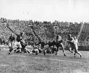 UM football vs Minnesota, 1937. A black and white image of the U-M football team playing Minnesota. Surrounded by a full stadium, players are tackling one another.