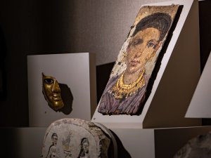 A case in the Kelsey Museum’s Egyptian galleries displaying a painted mummy portrait of a woman wearing gold jewelry, a gold-painted fragment of a mummy mask, and two stelae carved with images of people and hieroglyphs.