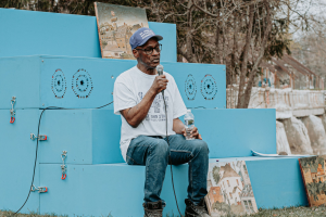 Photo of a man speaking into a microphone while sitting on a set of light blue steps (created by ARIA awardee UM-Flint Assistant Professor of Art Ash Arder), taken during the St. John Street Neighborhood block party (part of a project led by ARIA awardee UM-Flint Associate Professor Benjamin Gaydos)