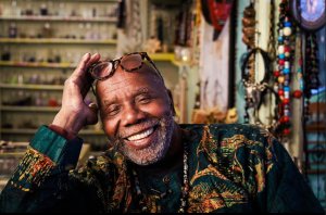 A Black man sits, laughing, in a studio filled with colorful beads