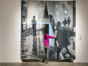 A man wearing a pink shirt walks through a beaded curtain created to show the images of Black figures