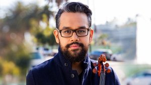 Vijay Gupta, a man with dark hair, a beard, and black-framed glasses, holds a violin