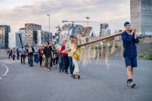A procession of people: the two leading it hold either end of a long wooden pole, draped with bundles of dried grasses.