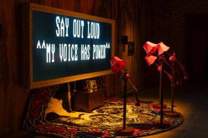 An interactive art installation featuring a large glowing screen that reads "SAY OUT LOUD **MY VOICE HAS POWER**" next to four red vintage floor lamps with numbered shades and telephone receivers.
