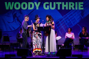 Three musicians perform with acoustic guitars on a purple-lit stage below large green text reading "WOODY GUTHRIE".