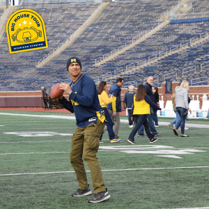 Man wearing a Michigan beanie and jersey gets ready to throw a football on the field as others in maize and blue walk around him at Michigan Stadium.