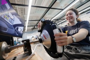 Hannah tests out a steering wheel with hands on its haptic feedback sensors while driving in a lab looking at the monitor.