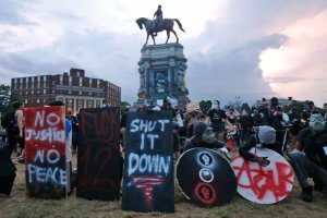 A crowd protests around an equestrian statue on a tall pedestal, many holding signs and wearing masks.