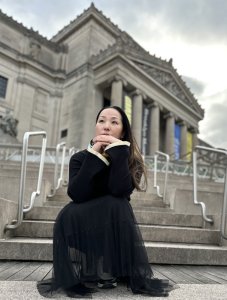 Kanako Uzawa sits outside of a large gray museum-like building, looking pensive up at the sky.