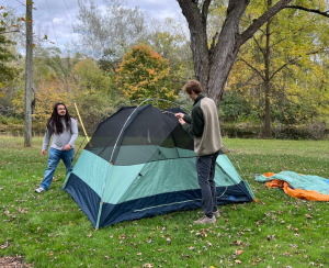 Students setting up a tent in a grassy area with trees.