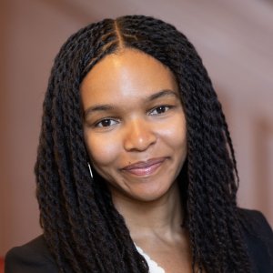 Headshot of a young Black woman with long dark twists parted down the center, smiling gently at the camera.