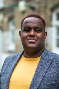 Headshot of Ismail Einashe wearing a gray blazer over a yellow shirt, standing outdoors against a softly blurred background.