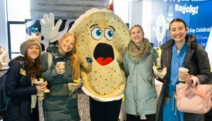 A group of students hold coffee and bagels while posing with the Alumni Association bagel mascot, Bagely