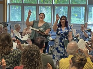 Two people standing with one arm raised leading a group of singers in performance from The Sacred Harp