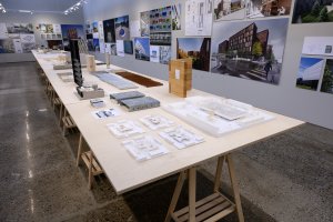 Photo of the exhibition space with images of the curator's work on the back wall and models on the long, central table.