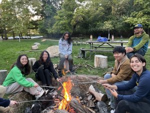 Smiling students are seated around a campfire in a wooded area