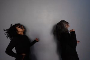 Two dancers attired in black long-sleeved shirts against a gray wall.