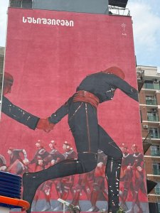 A mural painted on a windowless building surface about 8 stories tall. In the mural, people wearing black and red full-body uniforms hold hands, with a red background and white lettering at the top.