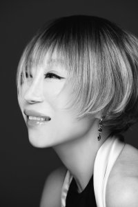 Alt Text for Accessibility:	A black-and-white portrait featuring a person in profile, turned slightly toward the viewer with a gentle smile that reveals their upper teeth. They wear a short, rounded bob haircut with piecey bangs falling near the eyes, light-toned layered strands framing the cheekbones, and dark winged eyeliner that accentuates the eyes. The look is styled with a dangling drop earring composed of geometric stones, a halter-style top featuring a light-colored neck strap over a dark garment, and soft studio lighting that highlights their features against a smooth, dark background.