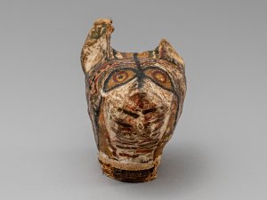 Head of a cat mummy made of stucco/plaster, missing one ear. Its highly decorated surface is painted with patterns in black, red, and yellow paint.