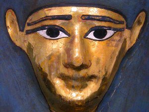 Close-up of a male Dynastic Egyptian mummy mask. It features straight eyebrows, kohl-lined eyes, and a long wig painted in blue-black, while the face itself is painted gold.