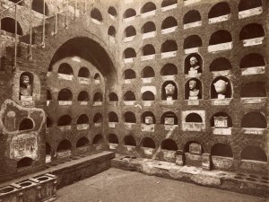 Sepia-toned archival photo of a Roman columbarium. A staircase leads into the space, which includes several rows of semicircular niches in the walls. Some of these recesses hold urns or statues of the deceased.