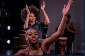 Two dancers with arms raised, and drummers in the background.