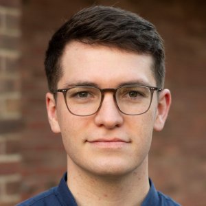 Headshot of a young white man with short brown hair, wearing dark-framed glasses and a blue shirt, looking directly at the camera against a soft, blurred background.