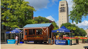 A wooden farm stand trailer sits in front of the belltower on S Ingalls mall with shelves filled with local produce and two tents on either side