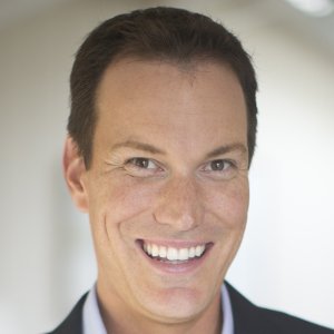 Headshot of Shawn Achor smiling, wearing a dark suit jacket over a light-colored collared shirt against a soft, out-of-focus background.