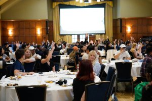 Photograph of students sitting at round tables in the League Ballroom. Several students are raising their hands to participate in an activity.