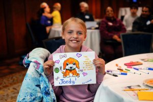 A young girl holds up an illustrated sign she made that says "my voice would be a golden retriever if it were an animal"
