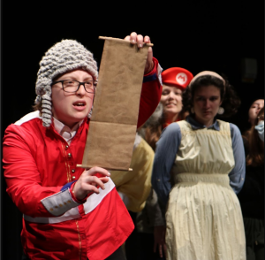 Several actors in costume onstage, one reading from a scroll while wearing a gray wig. The actors in the background are out of focus, two of the actors in view wearing a dress with a collar and a Mario hat.