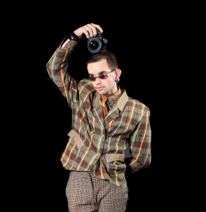 William Hohe, wearing round sunglasses and a plaid jacket, holds a camera above their head