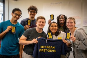 Six students standing in a line holding stickers that have a first-gen flag and two students are holding a Michigan First-Gens t-shirt in the First-Generation Gateway