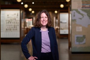 Portrait of Susanne Paulus, Associate Professor of Assyriology and Curator of the Tablet Collection at the University of Chicago’s Institute for the Study of Ancient Cultures. She smiles at the camera in a museum gallery, wearing a navy blazer over a lavender shirt, with softly blurred exhibit cases and maps behind her.