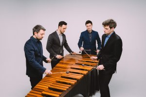 Four men wearing dark suits are gathered around and playing a xylophone simultaneously