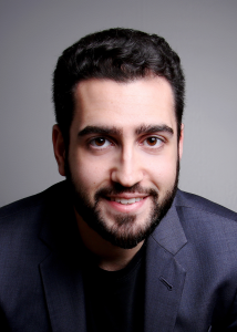 Headshot of Daniel Ohanian smiling at the camera, wearing a dark blue blazer over a black shirt against a neutral gray background