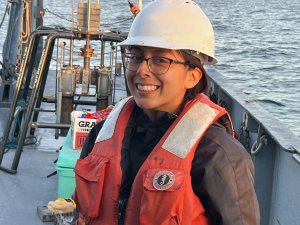 A woman in a hard hat and life jacket smiles at the camera from the deck of a boat outfitted with scientific equipment