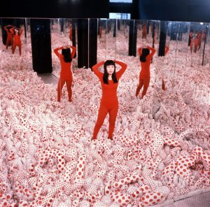 A 1965 photo of Kusama Yayoi standing in one of her exhibits. She is wearing a red bodysuit and standing among white and red polka-dotted 3-D forms, and there are mirrors behind her.