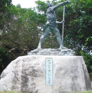 A photo of a statue on a rock outdoors. The statue is a realistic depiction of Oyake Akahachi carved from grey stone. The figure is shouting and pointing emphatically.