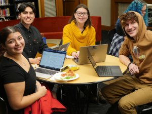 four smiling people sit at a round table and work on laptops