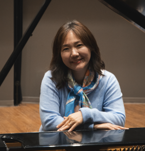 Lisa Sung, composer and pianist, smiles while seated at a grand piano. She wears a light blue sweater and a colorful patterned silk scarf, with her hands resting on the piano’s glossy black edge. The background features a wooden floor and dark structural beams in a music studio or rehearsal space.