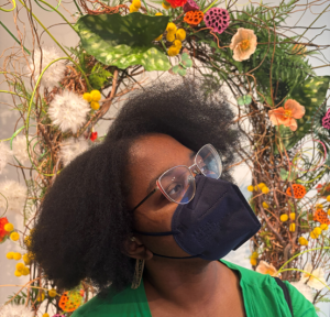 A female-presenting person in glasses and a black face mask looks to her left in front of a floral wreath that looks like a crown above her head