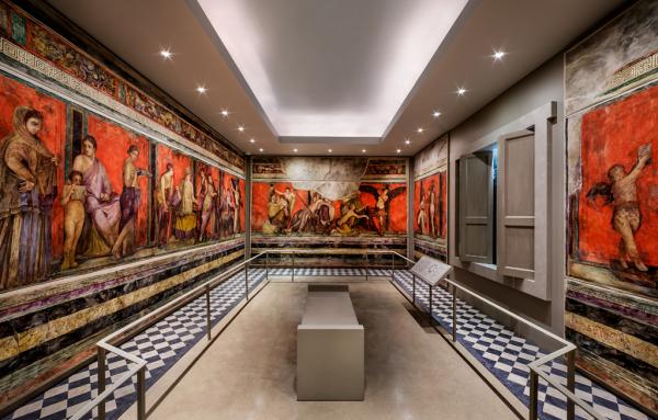 A rectangular room with a bench in the center. The walls are painted with an elaborate scene of men and women from Ancient Rome. Some of the people are mythological creatures and gods like cupids (boys with wings), maenads (nude women), and satyrs (men and boys with horse ears). The god Dionysus reclines against a seated woman's legs in the center back of the room while the women, maenads, and satyrs prepare for an event.
